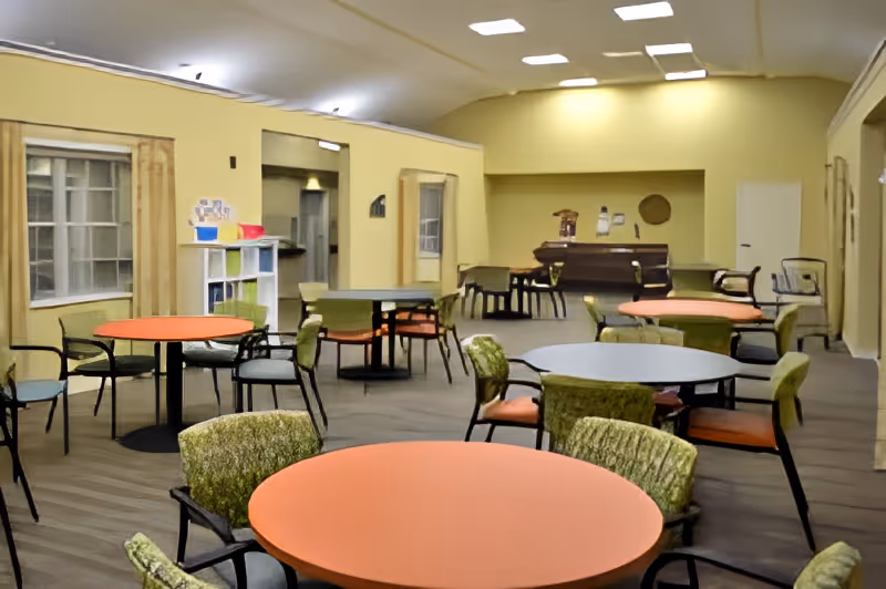 A spacious common area with multiple round tables and chairs arranged throughout the room. The walls are painted yellow, and there are windows with wooden frames on the left side. In the back of the room, there is a piano and some additional seating. The ceiling has recessed lighting, and the floor is covered with wood-like flooring.
