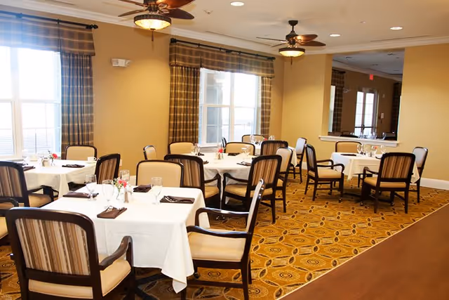 A dining room with several tables covered in white tablecloths, each set with glasses, napkins, and small flower arrangements. The room has large windows with plaid curtains, beige walls, patterned carpet, and ceiling fans with lights.