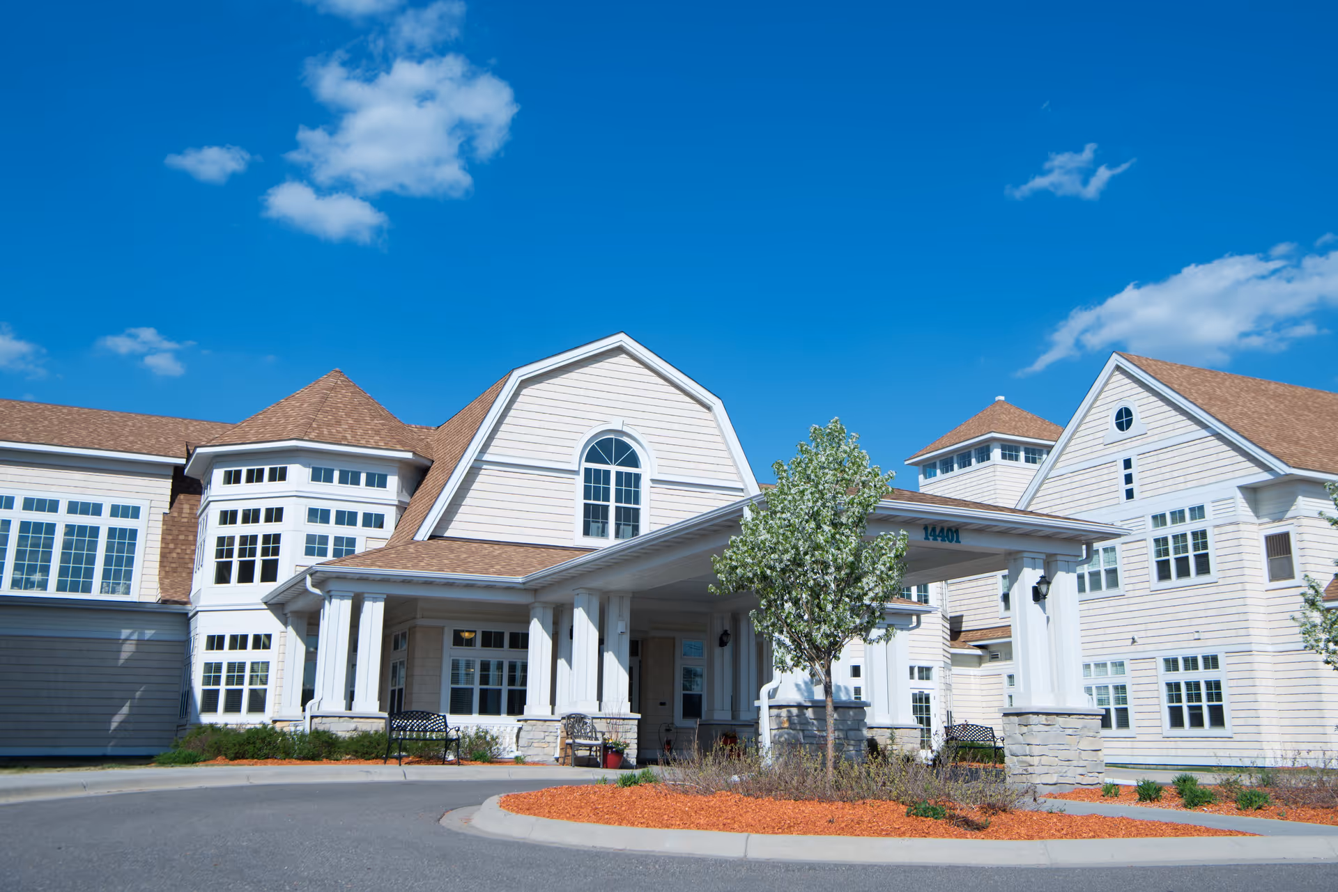 Exterior view of a large, two-story assisted living facility with white siding and brown shingled roofs under a bright blue sky with a few clouds. The building features multiple windows, a covered entrance with white columns, and landscaped areas with small trees and mulch.