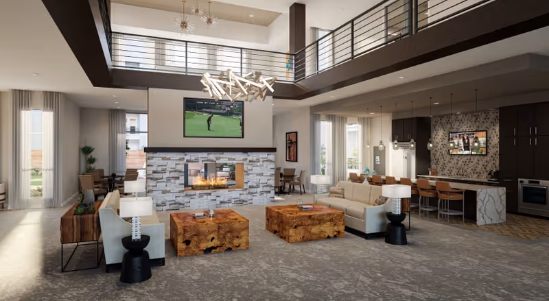Spacious and modern living room area with a double-sided stone fireplace in the center, two beige sofas facing each other, two wooden coffee tables, and a large flat-screen TV mounted above the fireplace. The room features high ceilings with a second-floor balcony railing visible, large windows with sheer curtains, and a kitchen area with a breakfast bar and stools in the background.