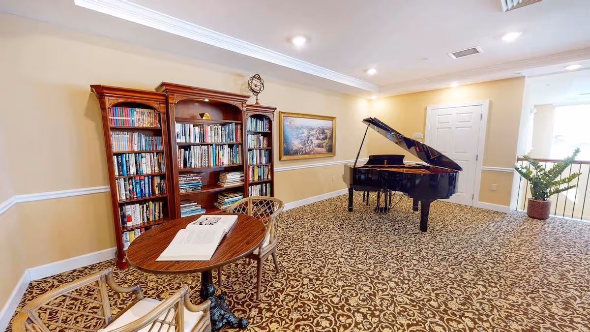 A cozy interior room featuring a grand piano, a wooden bookshelf filled with books, a small round table with an open book, and two chairs. The room has patterned carpet flooring, beige walls with white trim, a framed painting on the wall, and a potted plant near a railing.