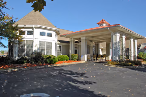 Exterior view of a senior living facility with a covered entrance supported by columns, a round turret-like section with large windows, landscaping with bushes and mulch, and a clear blue sky.