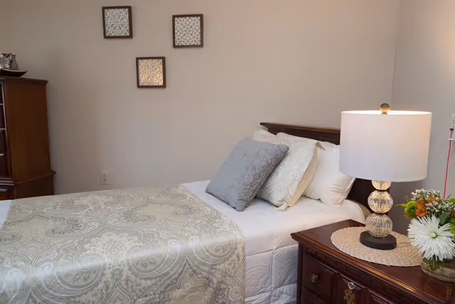 A neatly made bed with a patterned beige and white bedspread, several pillows including a gray decorative pillow, a wooden nightstand with a lamp and a small vase of flowers, and three framed decorative pieces hanging on a beige wall.