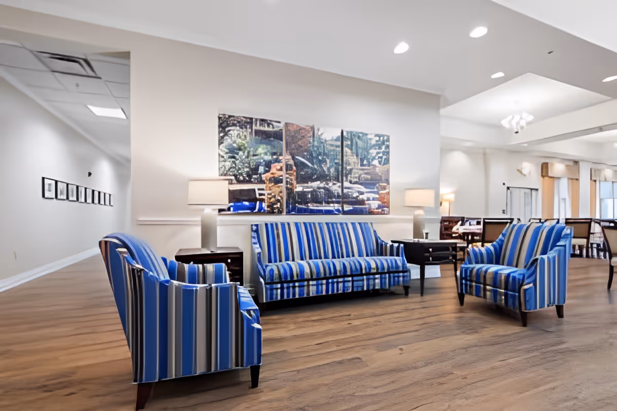 A bright and spacious living room area in Morada Deer Park featuring three blue and white striped upholstered chairs arranged around two dark wooden side tables with lamps. The room has wooden flooring, white walls, and a large triptych artwork hanging on the wall. In the background, there is a dining area with tables and chairs and large windows letting in natural light.