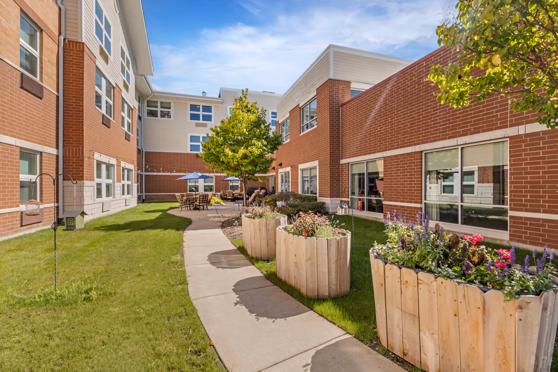 Outdoor courtyard area at The Sheridan at Tyler Creek featuring a curved concrete walkway, green grass, wooden planters with colorful flowers, a tree, and patio tables with umbrellas next to a red brick building under a blue sky with some clouds.