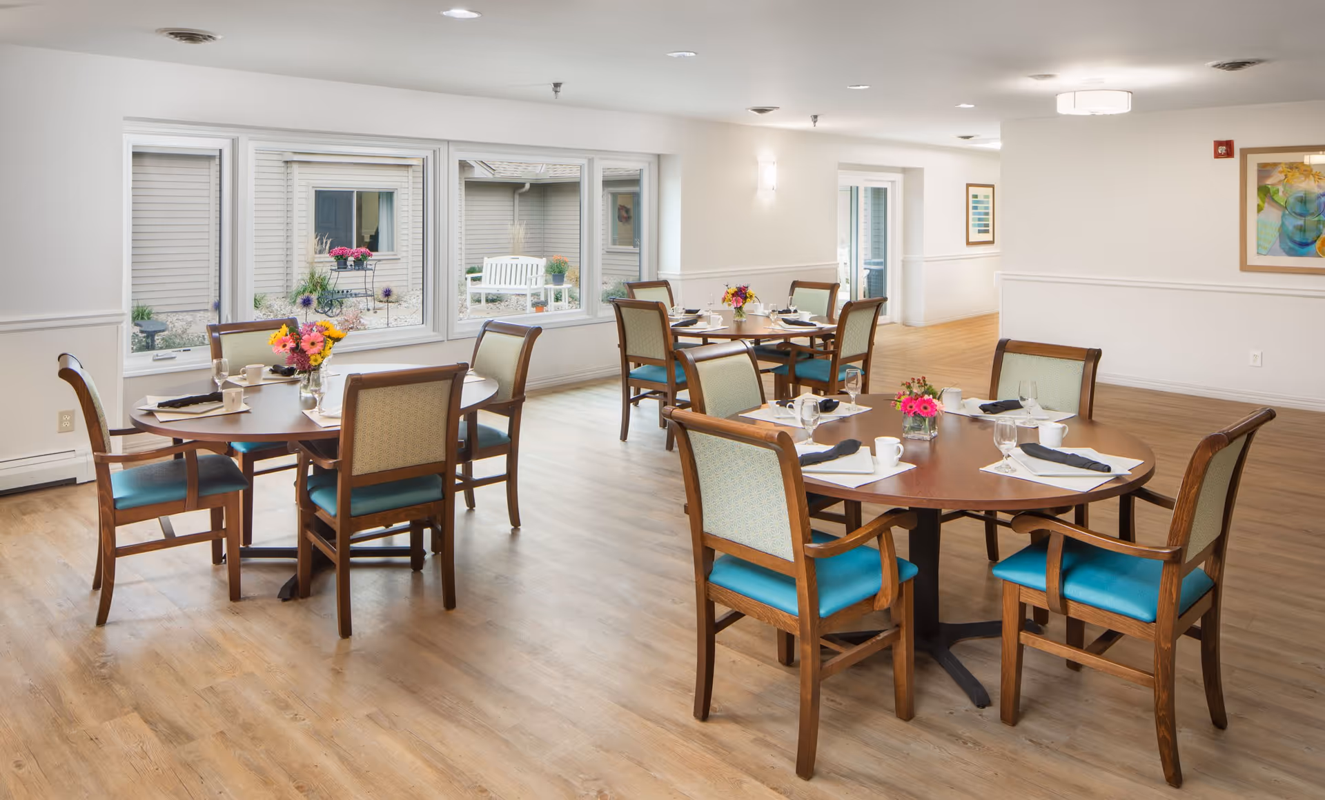 A bright dining room with round wooden tables and chairs with teal cushions. Each table is set with white cups, glasses, napkins, and small floral centerpieces. Large windows provide a view of an outdoor courtyard with a white bench and plants. The room has light-colored walls and wood flooring.