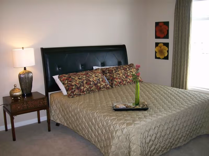 A bedroom with a large bed covered in a beige quilted bedspread and two patterned pillows. There is a wooden nightstand with a decorative lamp and a small golden container. On the bed is a tray with a green vase holding pink flowers. Two floral paintings hang on the wall next to a window with beige curtains.
