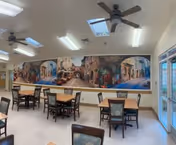 Interior view of a dining room with several wooden tables and chairs arranged neatly. The room has a large mural on one wall depicting a scenic street with buildings and flowers. The ceiling has skylights and ceiling fans, and there are large windows and glass doors on one side letting in natural light.