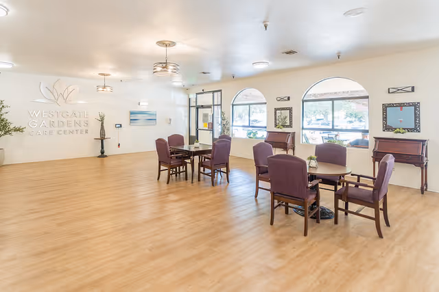 A spacious, well-lit common area in Westgate Gardens Care Center with wooden flooring, several tables surrounded by purple upholstered chairs, large arched windows letting in natural light, and decorative plants and artwork on the walls.