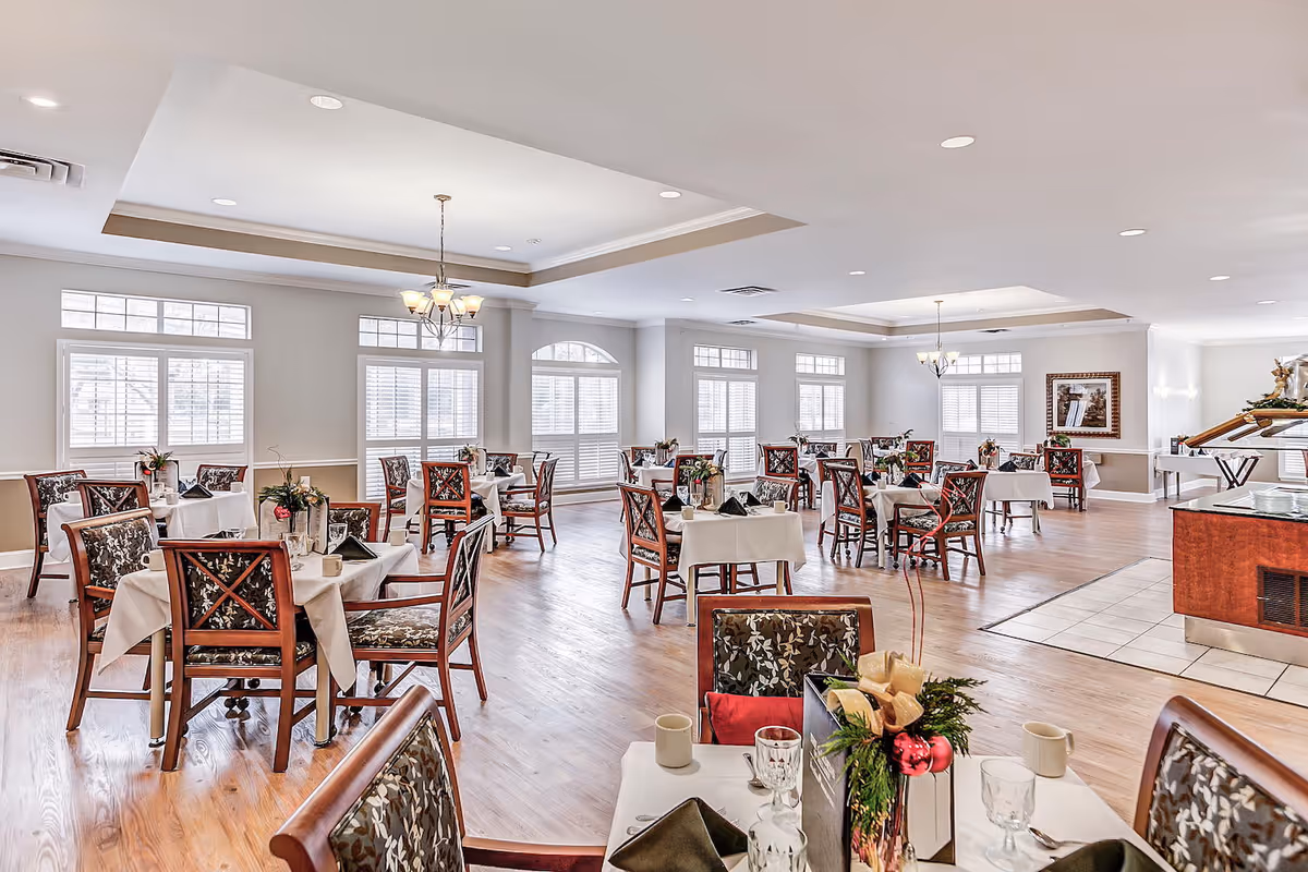 A spacious and well-lit dining room with multiple tables covered in white tablecloths, each set with glassware, napkins, and floral centerpieces. The room features large windows with white shutters, wooden chairs with patterned cushions, and chandeliers hanging from the ceiling. The floor is a combination of wood and tile near a buffet area.