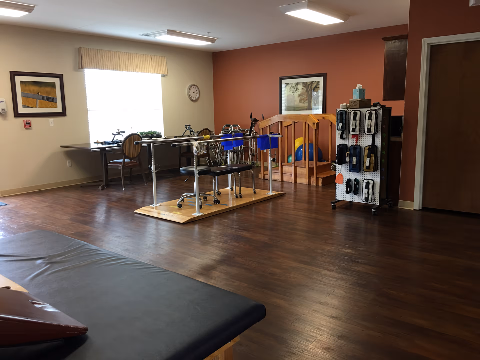 A rehabilitation room with wooden flooring featuring parallel bars for walking exercises, chairs, a table with two chairs near a window, a rack with various therapy equipment, and framed artwork on the walls.