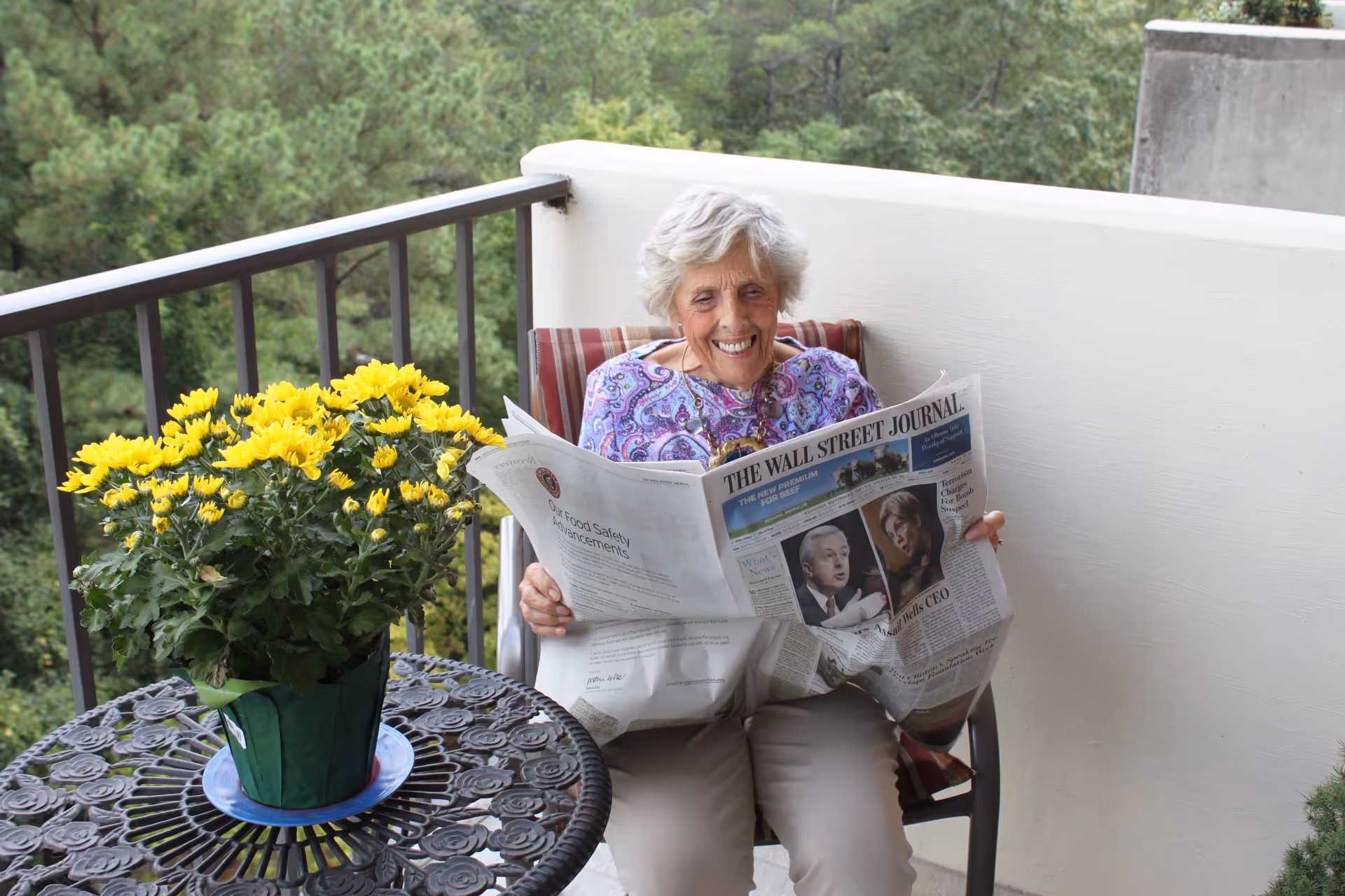 An elderly woman with gray hair sitting on a balcony chair, smiling while reading The Wall Street Journal newspaper. There is a round metal table with a pot of yellow flowers next to her, and green trees are visible in the background.