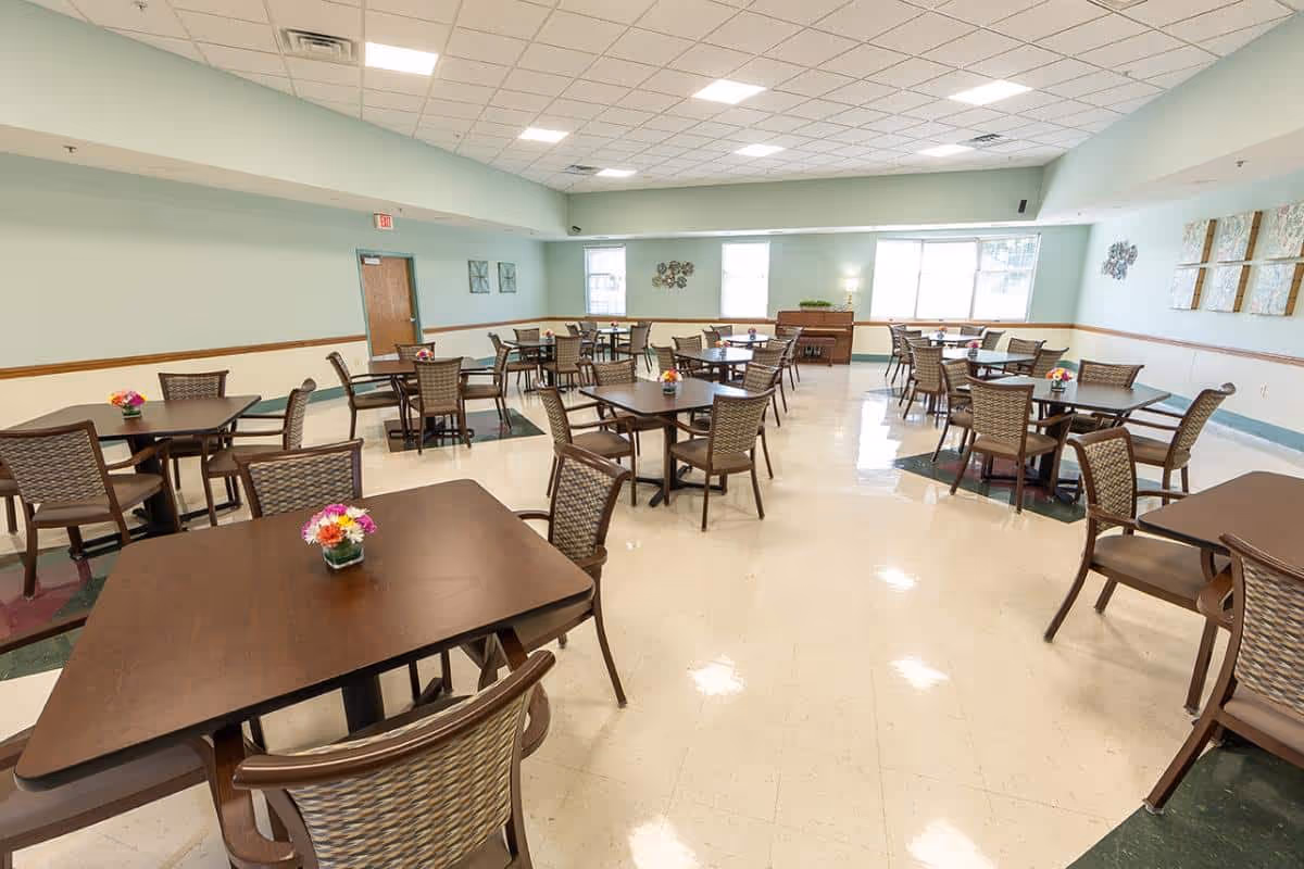 Bright, spacious dining room with multiple square tables and woven chairs arranged across the room, each table holding a small flower arrangement.