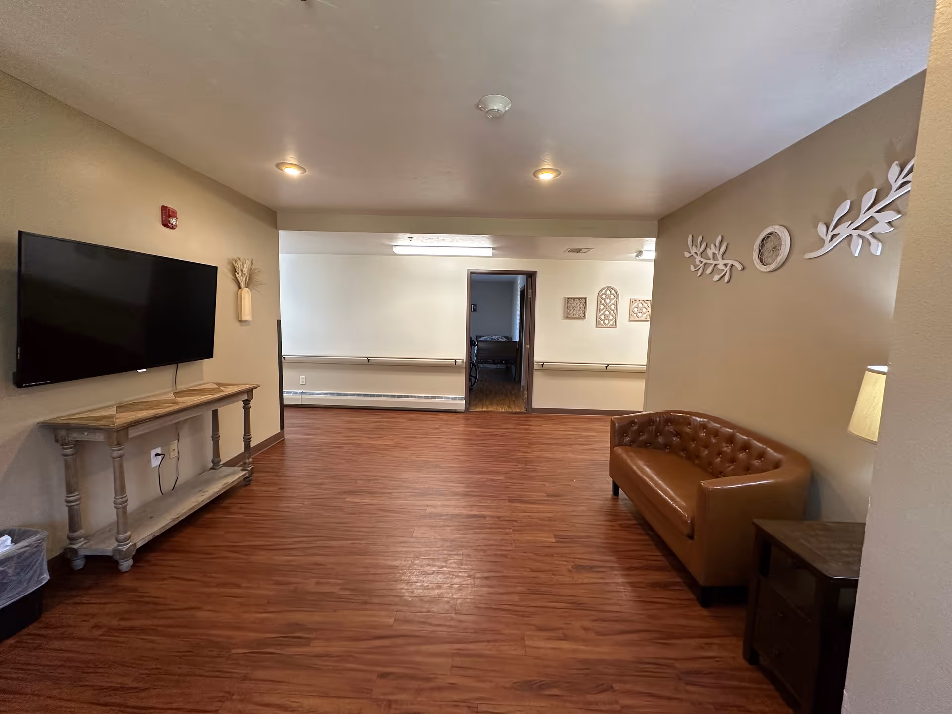 A room with wood flooring and beige walls featuring a mounted flat-screen TV on the left wall above a wooden console table. On the right side, there is a brown leather tufted sofa next to a small wooden side table with a lamp. The back wall has a doorway leading to another room and is decorated with wall art including white leaf designs and framed pieces. The ceiling has recessed lighting.