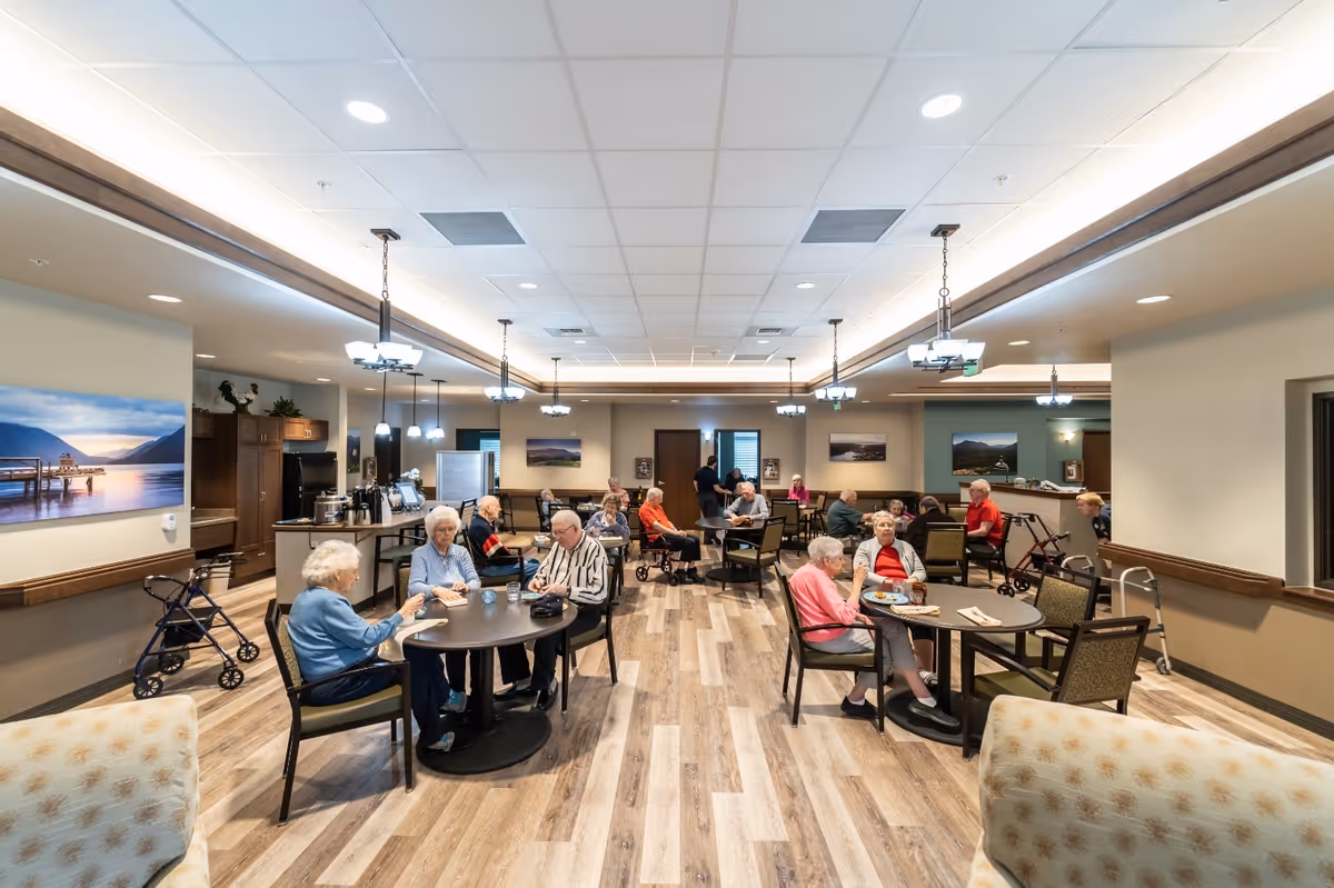 A spacious dining room in a senior living facility with several elderly residents sitting at round tables, eating and socializing. The room has wood-patterned flooring, modern ceiling lights, and landscape paintings on the walls. Walkers and mobility aids are visible near some residents.