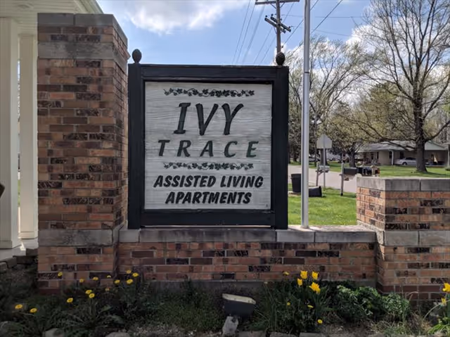 A brick sign at the entrance of a facility that reads 'IVY TRACE ASSISTED LIVING APARTMENTS' with some yellow flowers and greenery at the base, and a residential neighborhood visible in the background under a partly cloudy sky.
