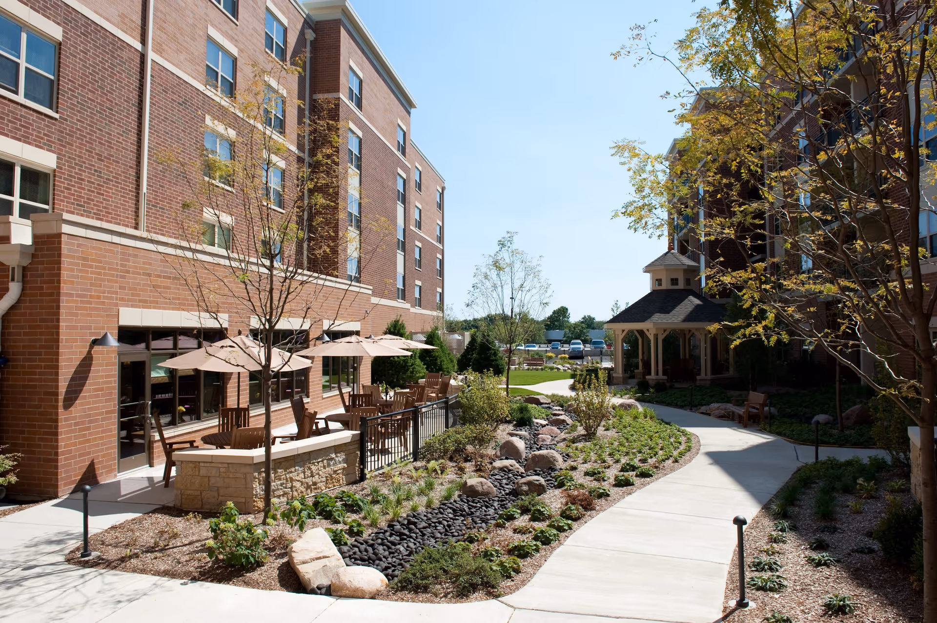 Outdoor courtyard area at Victory Centre of Vernon Hills featuring a paved walkway, landscaped garden with rocks and plants, wooden tables with umbrellas, and a gazebo. The courtyard is surrounded by multi-story brick buildings under a clear blue sky.