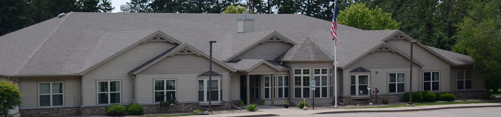 Single-story beige assisted-living building with a gabled roof, central entrance with a flagpole, and low landscaping in front.