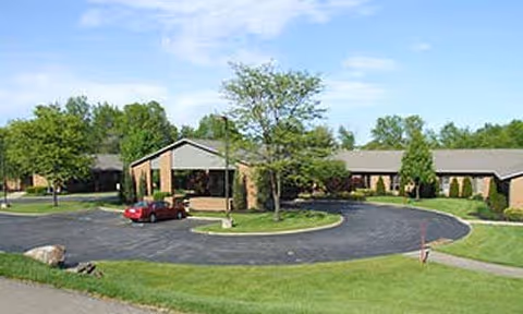 Single-story brick senior living facility with circular driveway, green lawns, and trees.