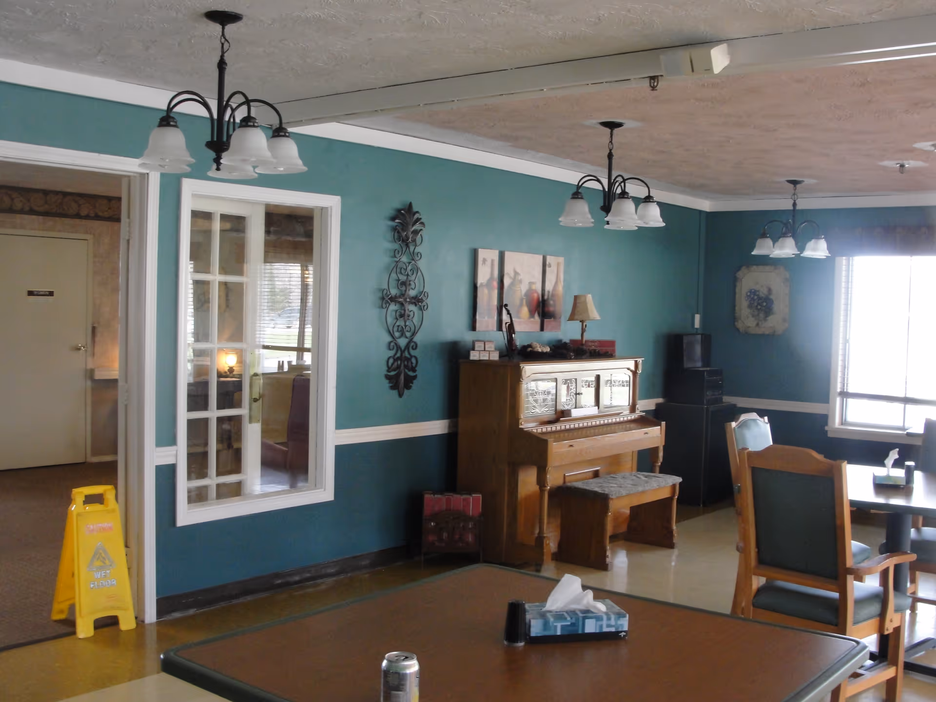 A senior living common area with teal walls featuring a piano, tables and chairs, hanging light fixtures, and a wet-floor sign.