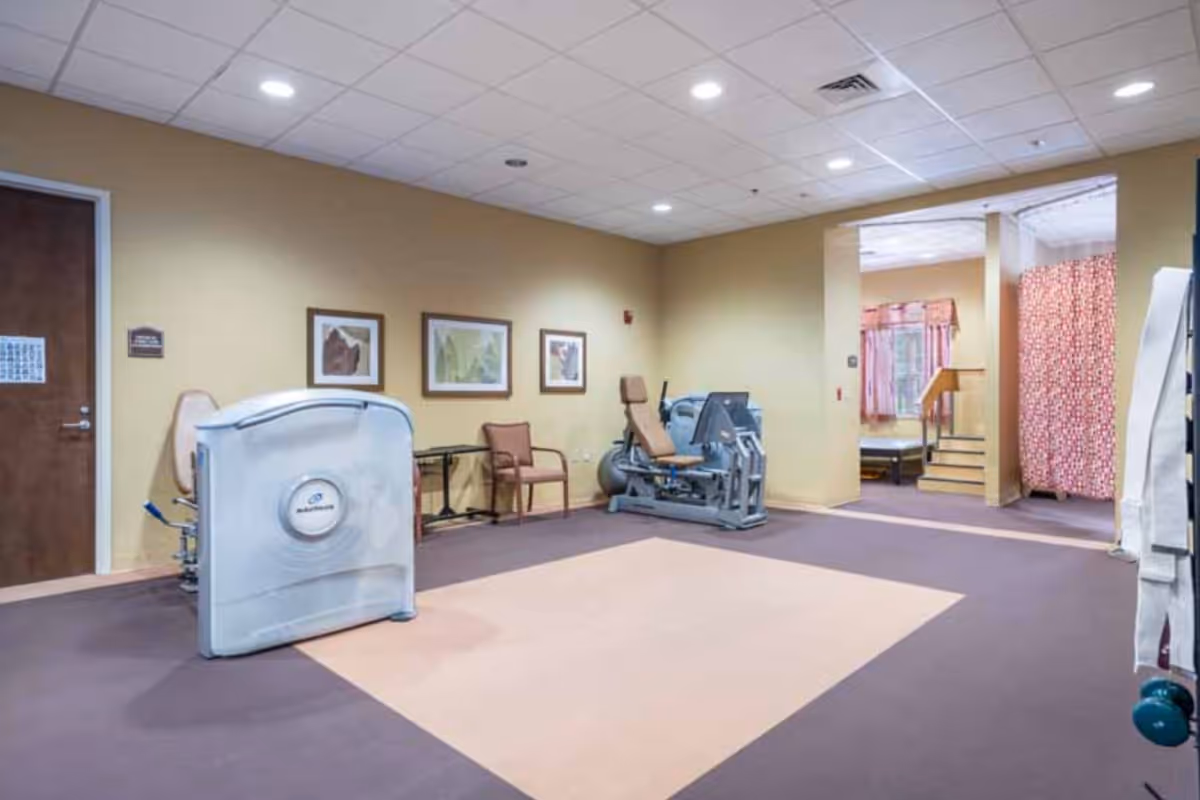 A rehabilitation or physical therapy room with exercise equipment, chairs, framed artwork on the walls, and a small raised platform with stairs and a handrail in the background.