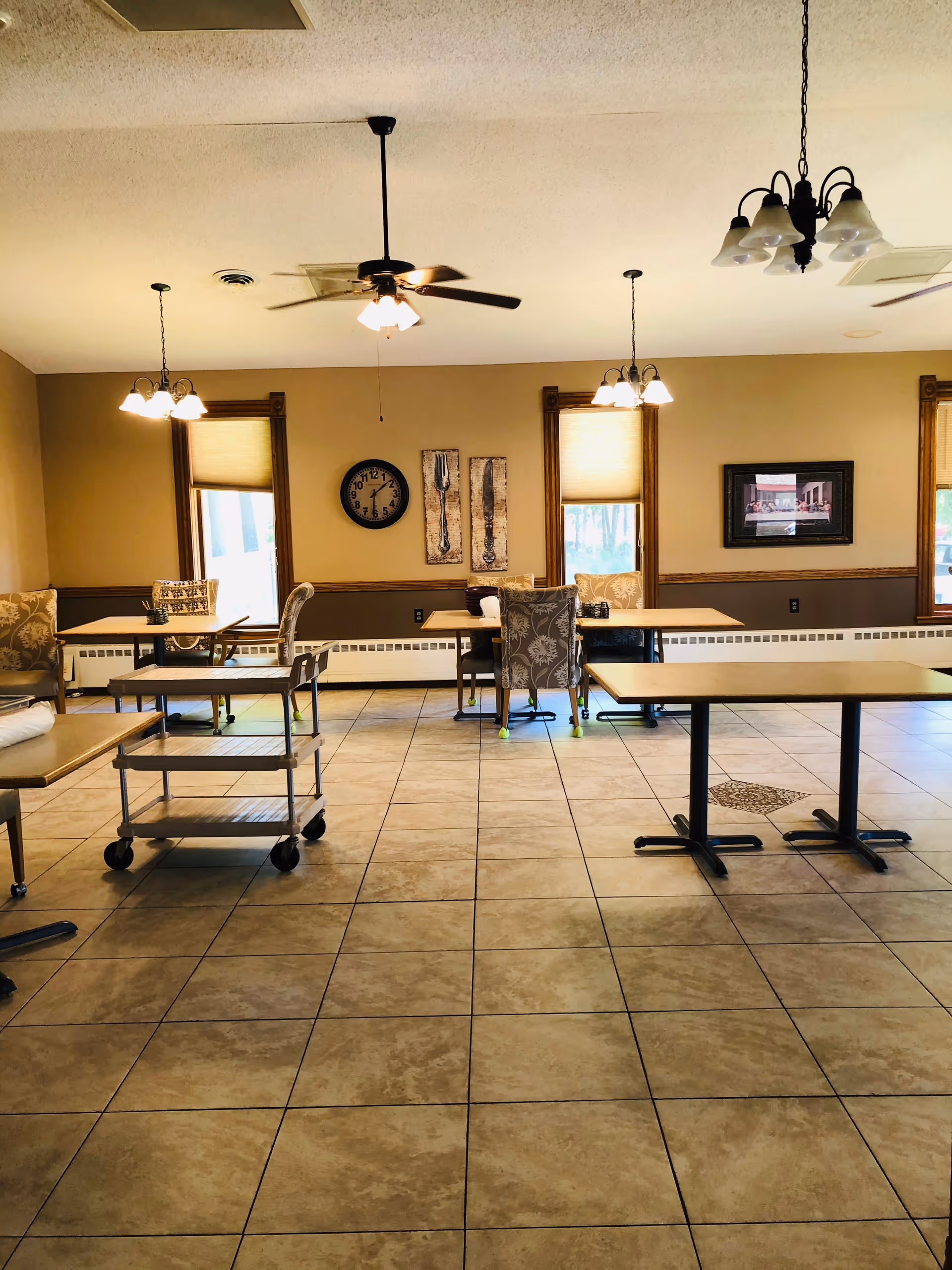 A spacious dining room with tiled floors, several tables and chairs arranged around the room, ceiling fans and hanging light fixtures. The walls are beige with wooden trim around the windows, which have shades partially drawn. There is a clock and decorative wall art featuring utensils on the far wall, along with a framed picture.