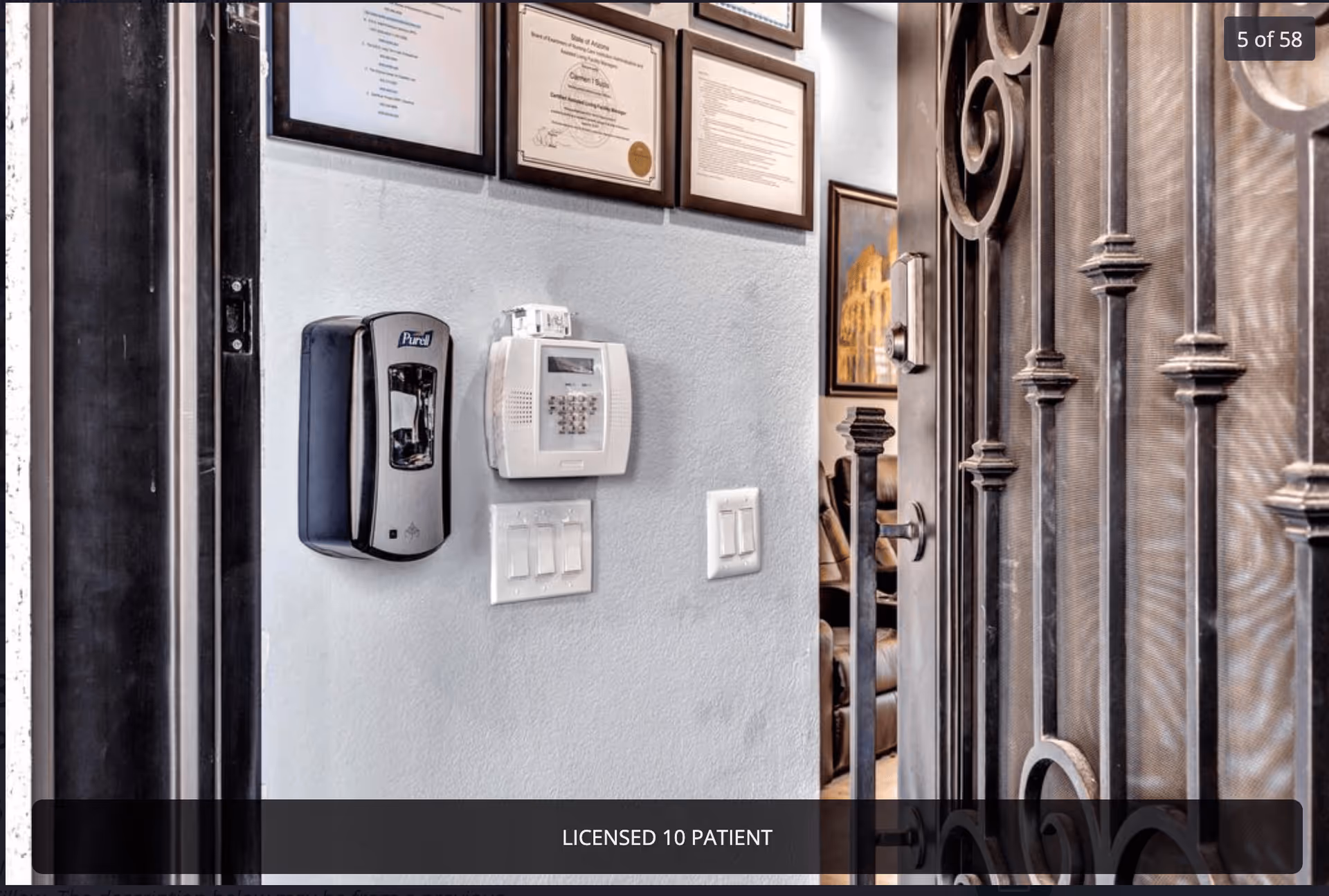 A close-up view of a wall inside a facility showing a Purell hand sanitizer dispenser, a security keypad, and multiple light switches. Above these are framed certificates and documents. To the right, there is a decorative metal door partially open, revealing a glimpse of a room with a leather chair and framed artwork on the wall.