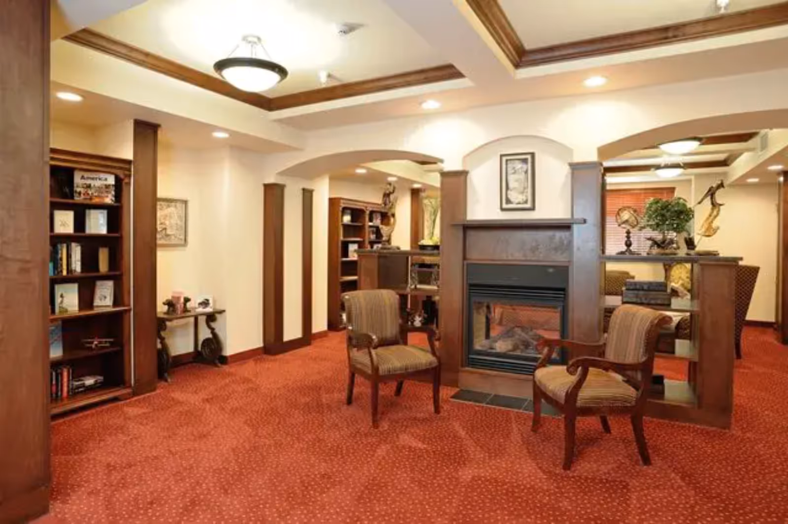 A cozy interior lounge area with two upholstered armchairs facing a wooden fireplace. The room features red carpet with a subtle pattern, wooden trim and columns, built-in bookshelves filled with books and decorative items, and warm lighting from ceiling fixtures.