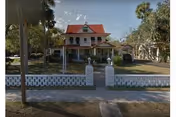 Two-story Victorian-style house with a red roof, wraparound porch and white columns, set behind a low decorative fence and palm trees.