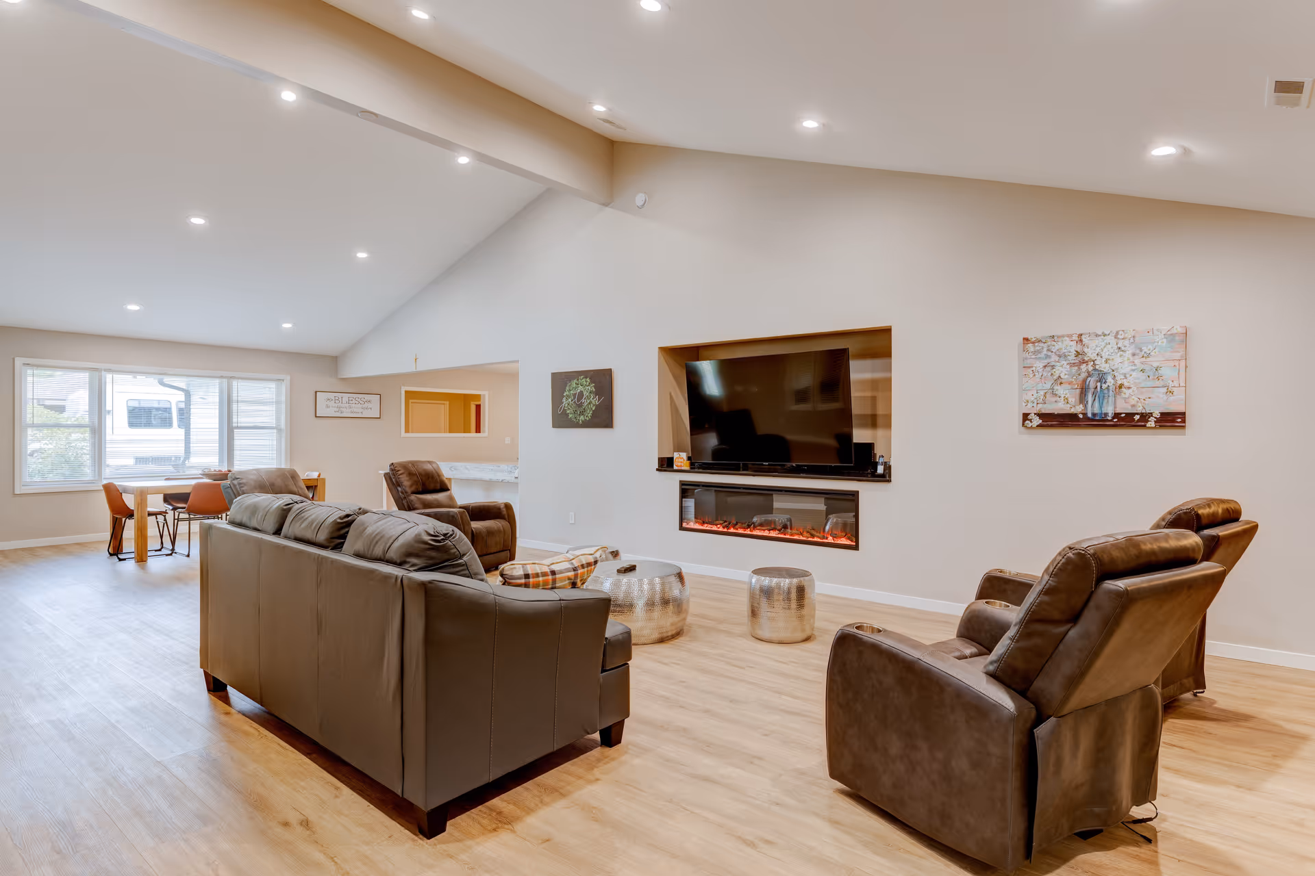 A spacious senior living room with a leather sofa, recliners, a wall-mounted TV above an electric fireplace, and a dining area in the background.