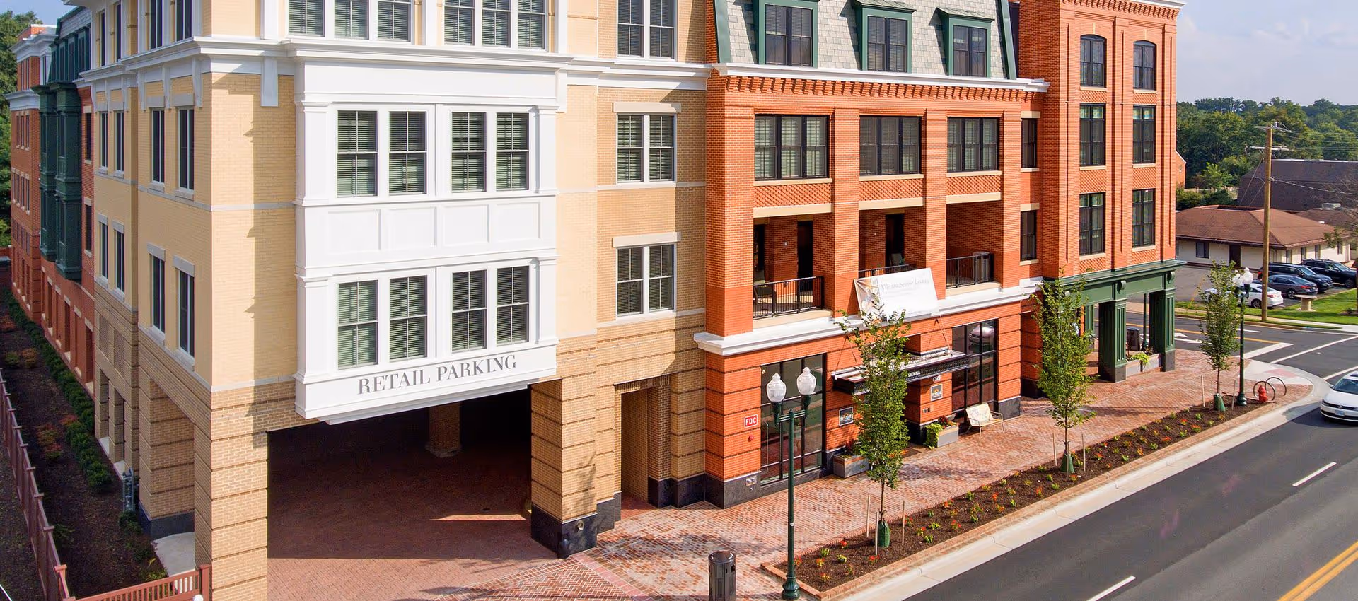 Exterior view of a multi-story building with retail parking entrance, brick and beige facade, street lamps, young trees, and a sidewalk along a road with parked cars.