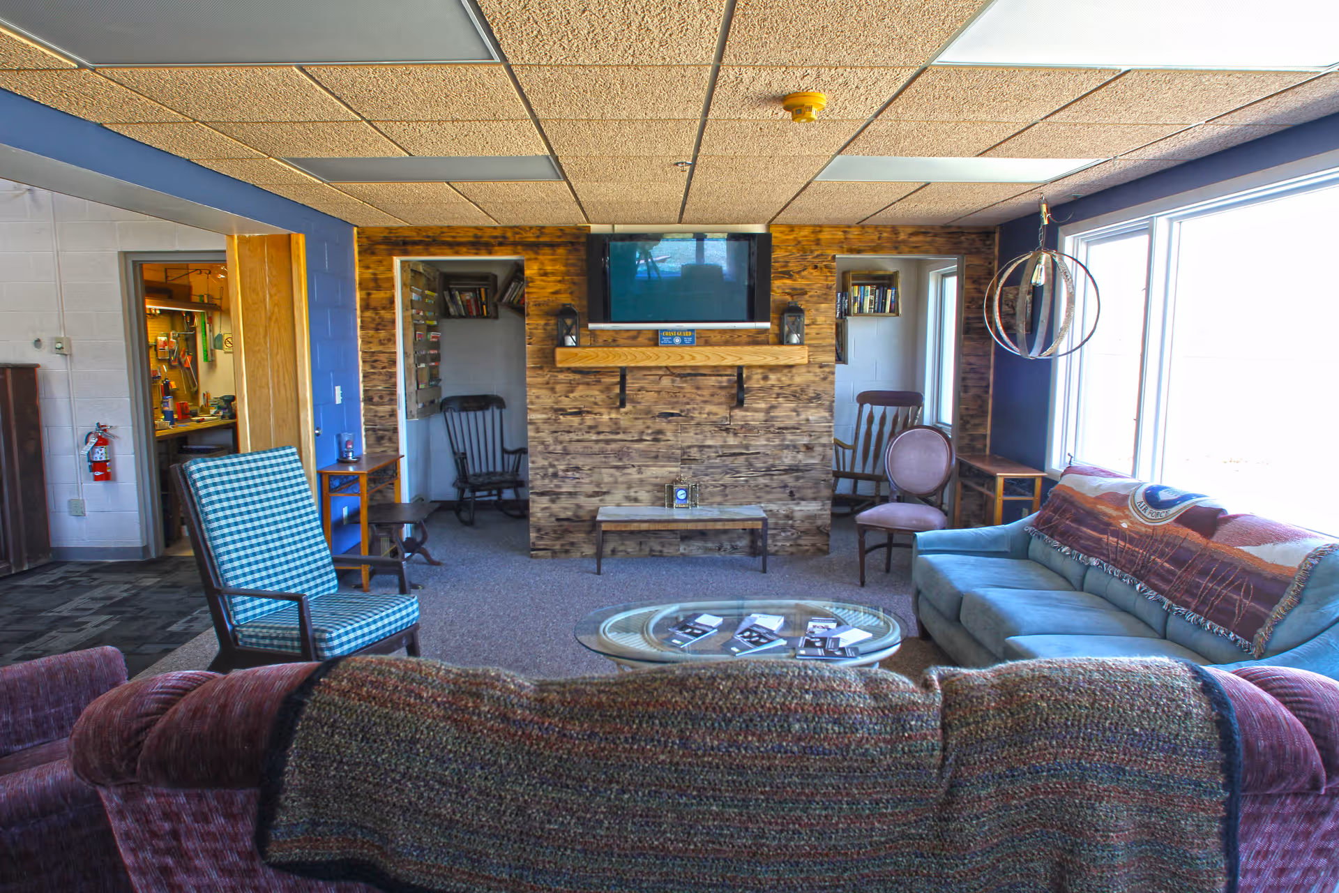 A cozy senior community living room with a variety of seating including a blue sofa, a patterned armchair, and wooden chairs. A rustic wooden wall features a mounted flat-screen TV and a small shelf. Large windows allow natural light to fill the room, and a round glass coffee table holds several pamphlets. The room has a carpeted floor and a drop ceiling with fluorescent lights.