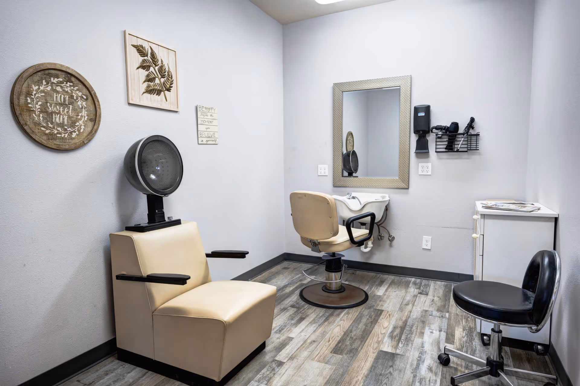 Interior view of a small salon room with a beige salon chair under a hair dryer, a beige salon chair in front of a white sink with a mirror above it, a black rolling stool, a white cabinet with magazines on top, and wall decorations including a round wooden sign that says 'Home Sweet Home' and framed leaf art.