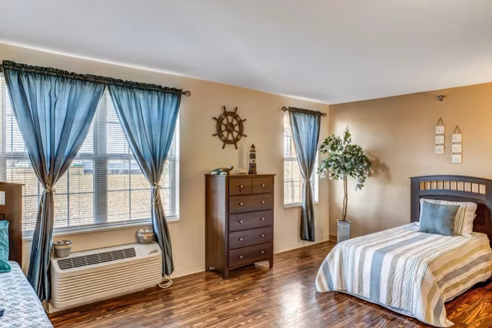 Well-lit bedroom with a single bed, wooden dresser, blue curtains, and a potted plant.
