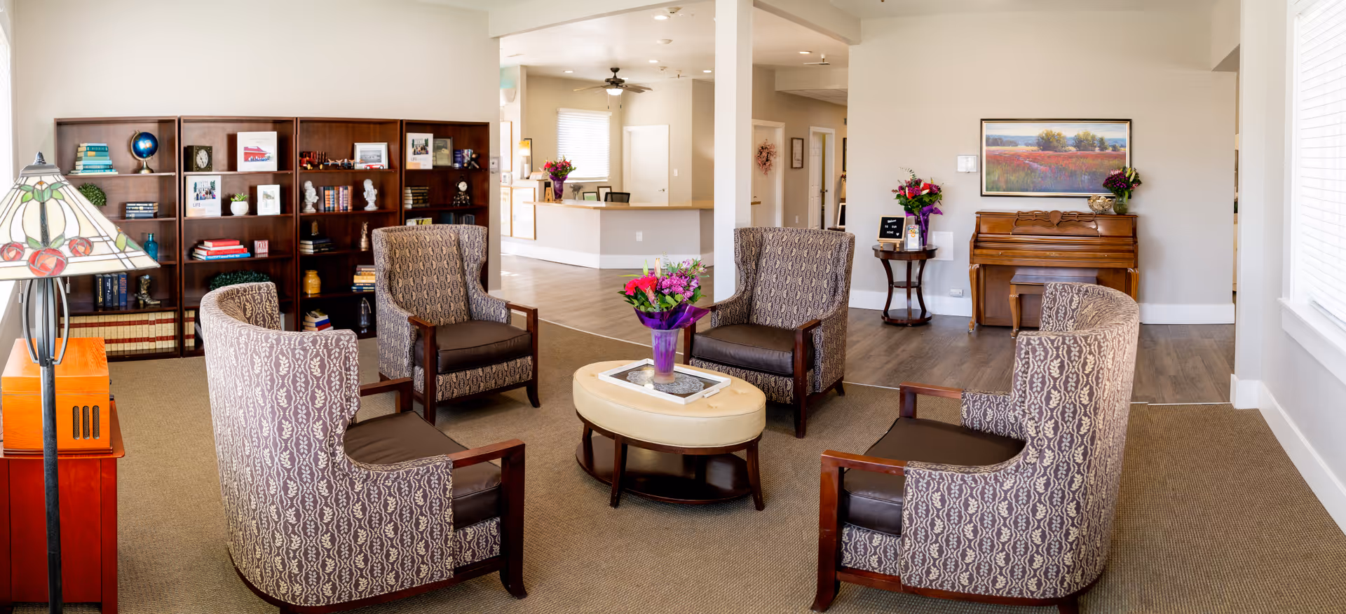 A cozy senior living common area with four patterned armchairs arranged around an oval ottoman with a flower vase on top. Behind the seating area is a wooden bookshelf filled with books and decorative items. To the right, there is a wooden piano with a framed landscape painting above it and a small round table with flowers. The background shows a reception desk and hallway with natural light coming through windows.
