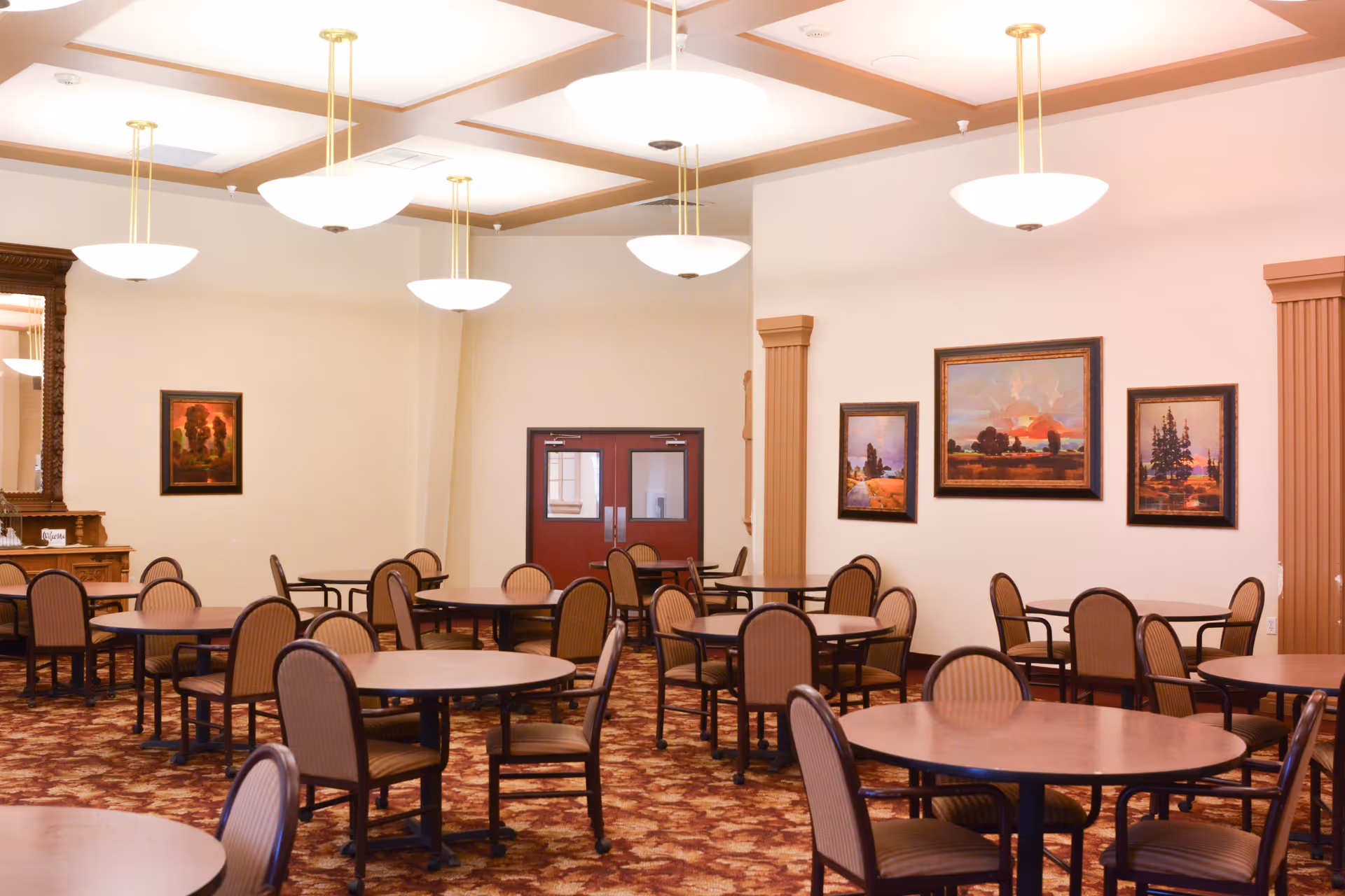 A spacious dining room with multiple round tables and chairs, framed landscape paintings, and hanging pendant lights.