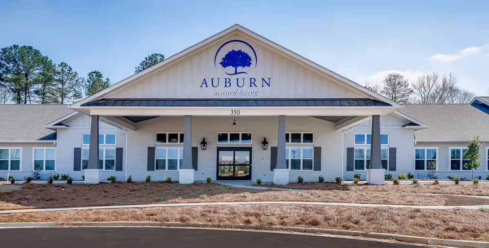 Front exterior view of Auburn Assisted Living facility, a single-story building with white brick walls, multiple windows with shutters, a covered entrance supported by columns, and a large sign with a tree logo and the name 'Auburn assisted living' above the entrance.