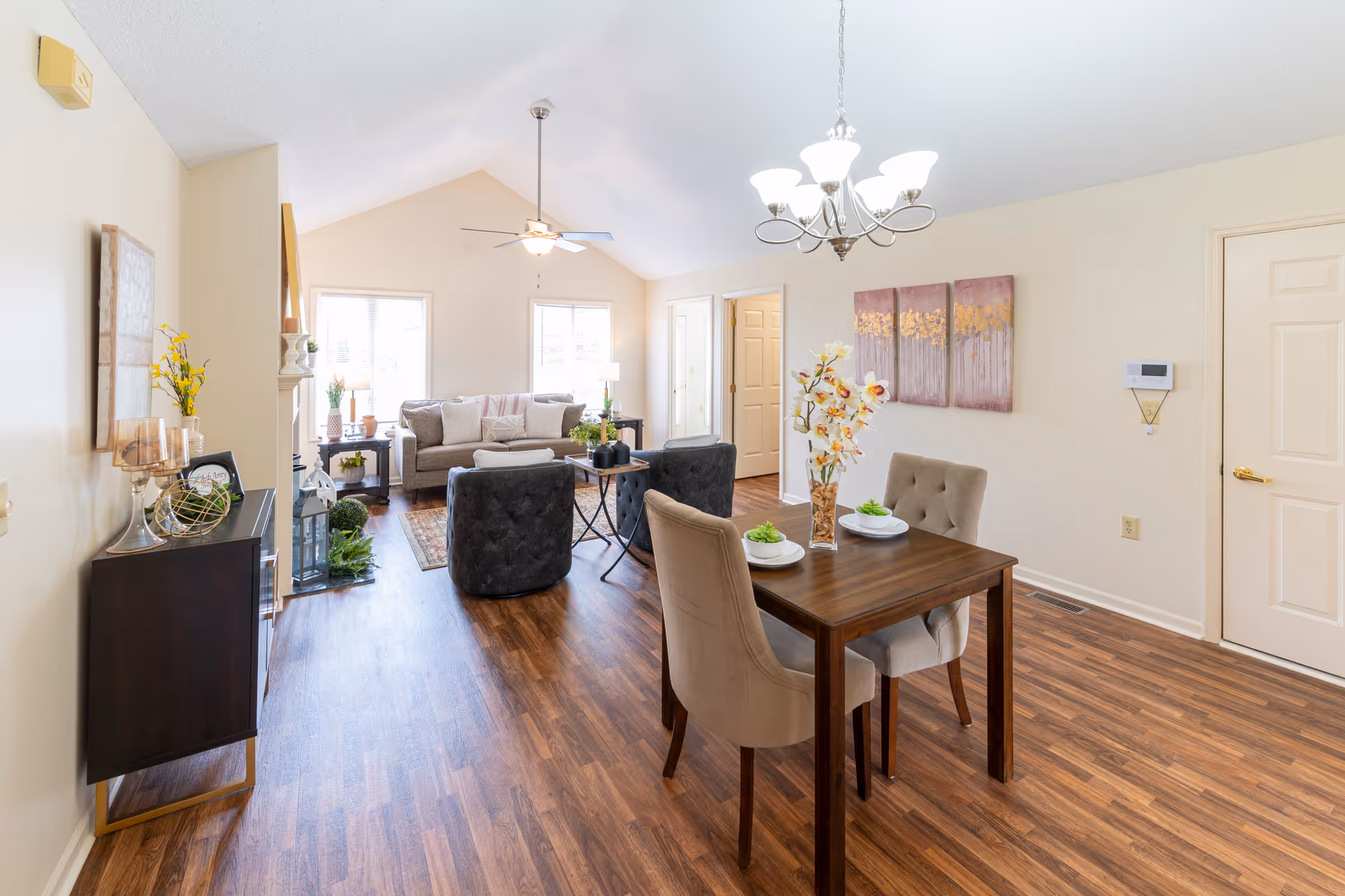 Bright and spacious senior living community interior featuring a dining area with a wooden table set for two, a chandelier overhead, and a living room with a sofa, two armchairs, side tables, and decorative plants. The room has wood flooring, light-colored walls, and large windows allowing natural light to fill the space.