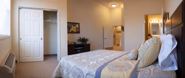 Sunlit bedroom with a patterned bed and pillows, an open closet, dresser, and a doorway leading to a bathroom.