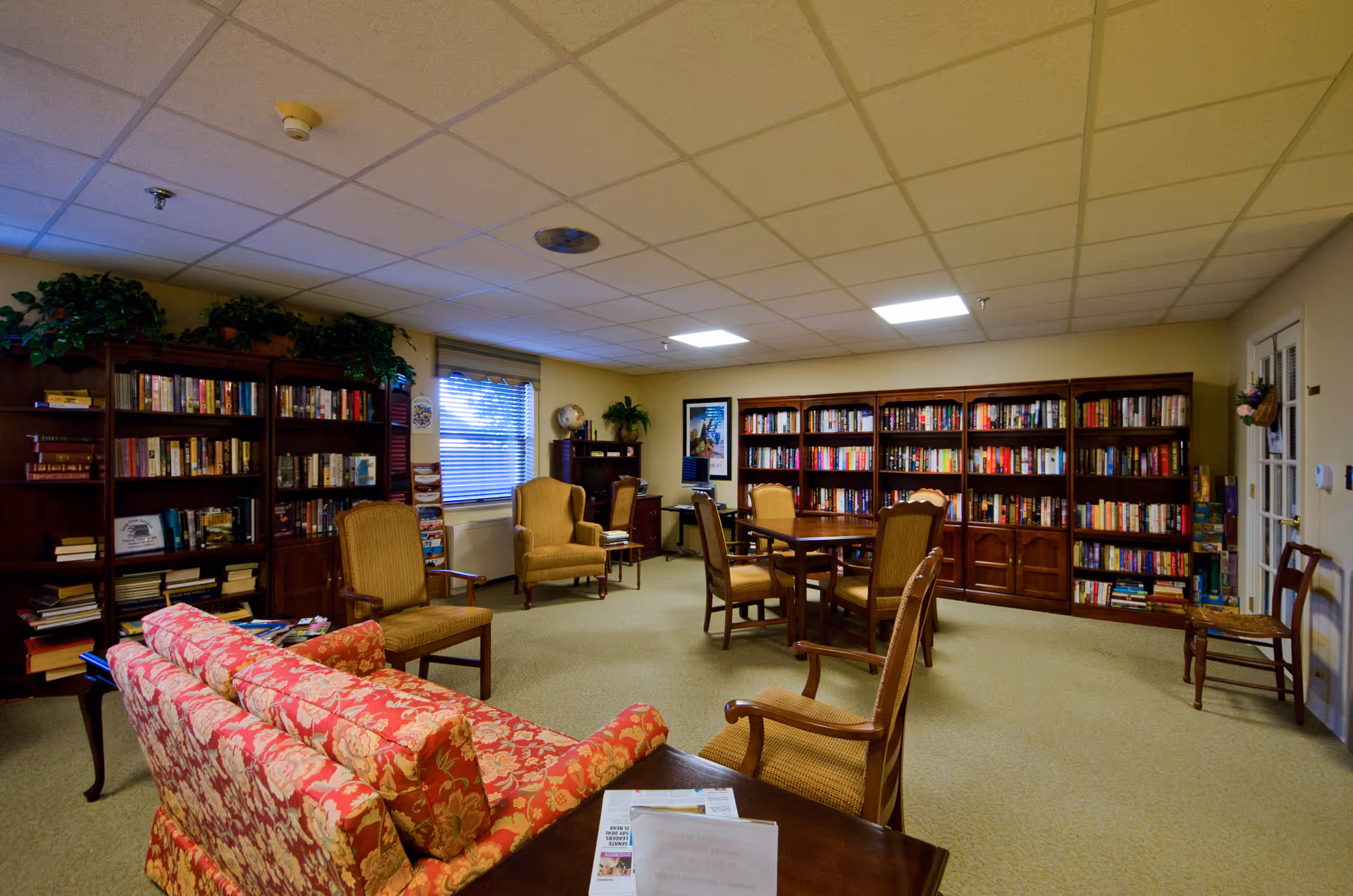 Communal library-style living room with bookshelves lining the walls, a floral sofa, and several armchairs and a table.