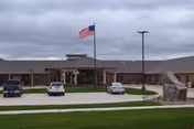 Single-story senior living community building with a covered main entrance, an American flag on a flagpole, several parked cars, and a landscaped front lawn with a stone sign.