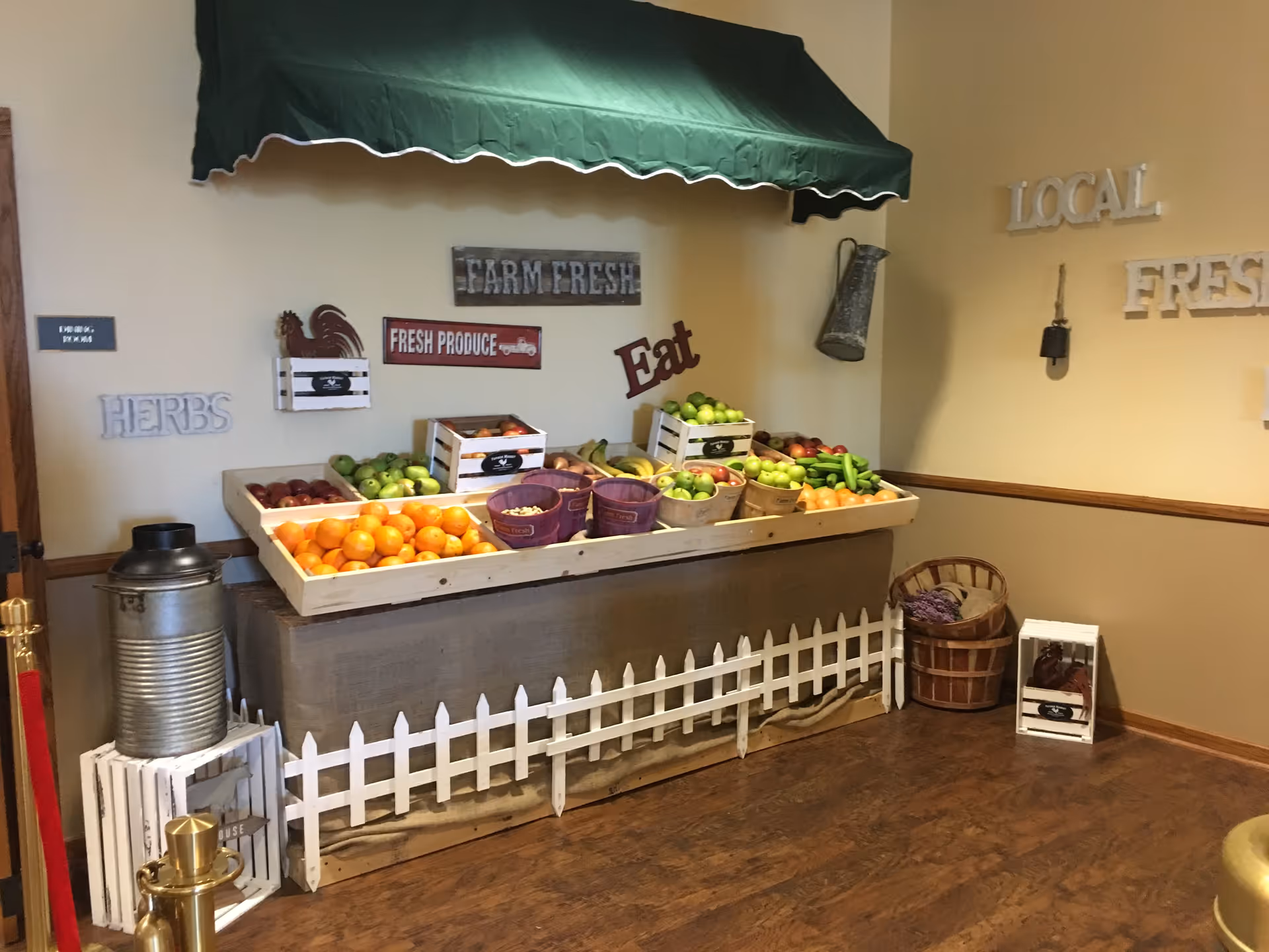 Indoor display of fresh produce including apples, oranges, bananas, and cucumbers arranged in wooden crates and baskets under a green awning with decorative signs on the wall reading 'Farm Fresh', 'Fresh Produce', 'Eat', 'Herbs', and 'Local Fresh'. The floor is wooden and there is a small white picket fence in front of the display.