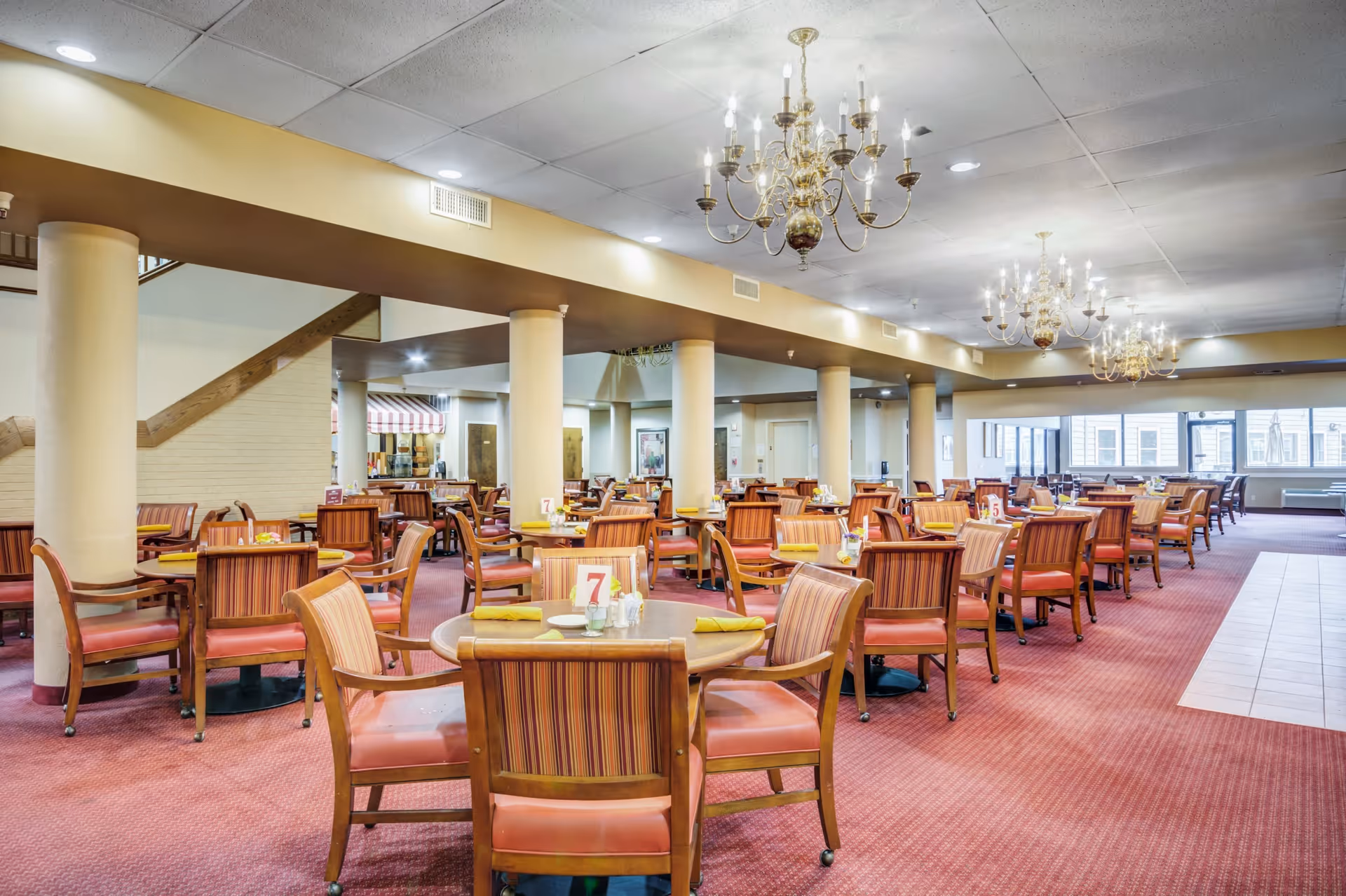 Spacious dining room in a retirement community with multiple round tables and wooden chairs with striped upholstery. The room features red carpeting, beige walls, large windows allowing natural light, and elegant chandeliers hanging from the ceiling.