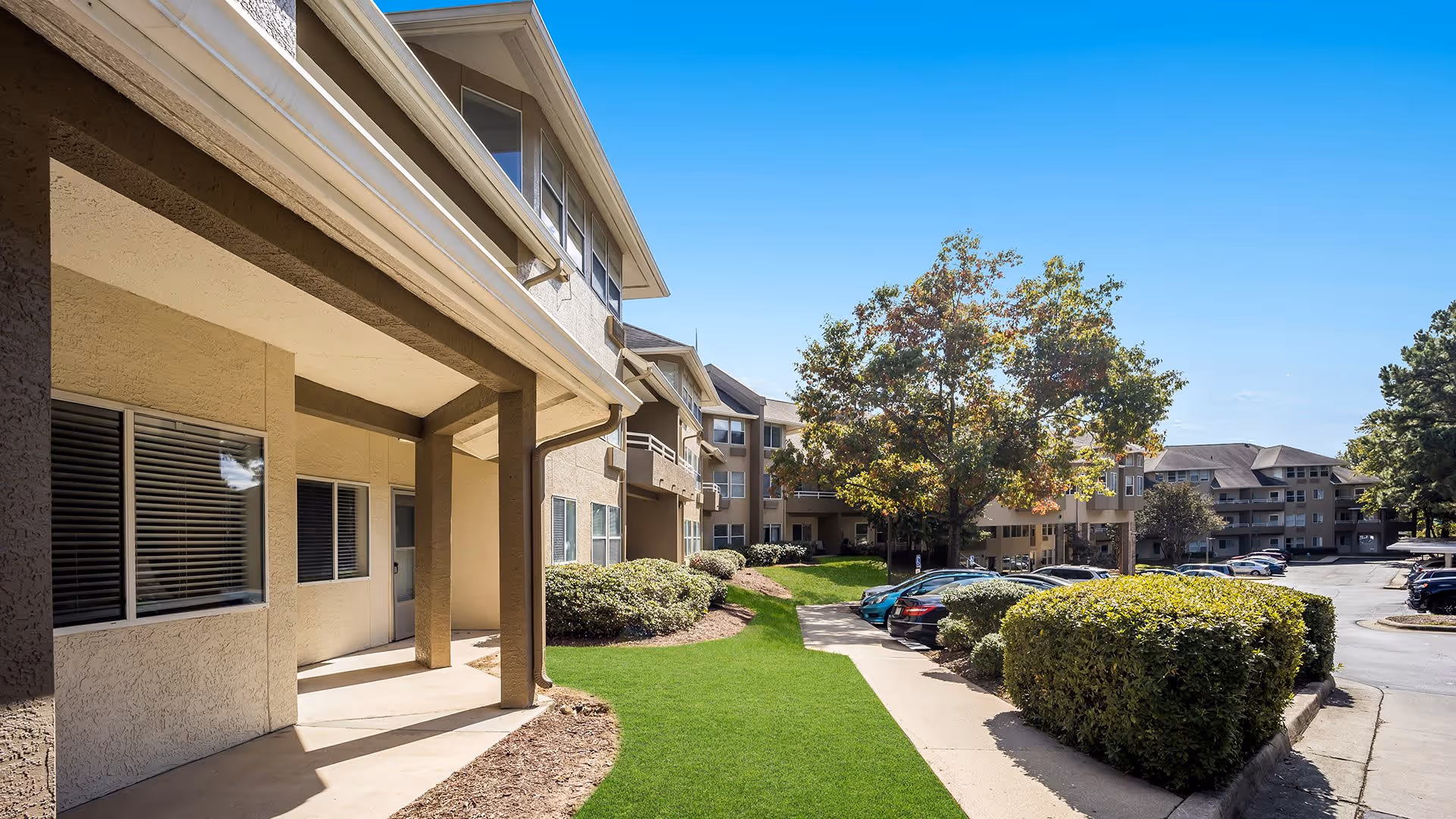 Exterior view of Holiday Durham Regent senior living facility showing a beige multi-story building with windows and balconies, a well-maintained green lawn, bushes, trees, a sidewalk, and a parking lot with several parked cars under a clear blue sky.