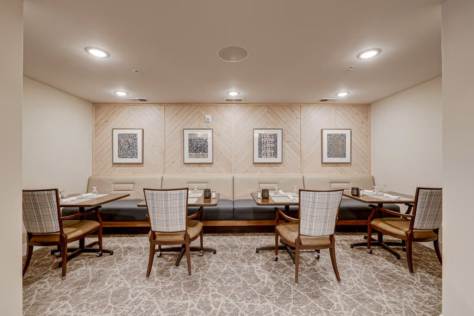 A dining area with four tables, each set with two chairs and a bench along the back wall. The tables have place settings with napkins and glasses. The wall behind the bench features light wood paneling with four framed abstract artworks. The floor is carpeted with a patterned design, and the ceiling has recessed lighting.