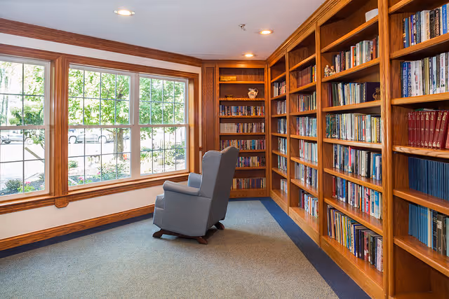 A cozy interior library with wooden built-in bookshelves, a single upholstered armchair, and a large window overlooking trees.