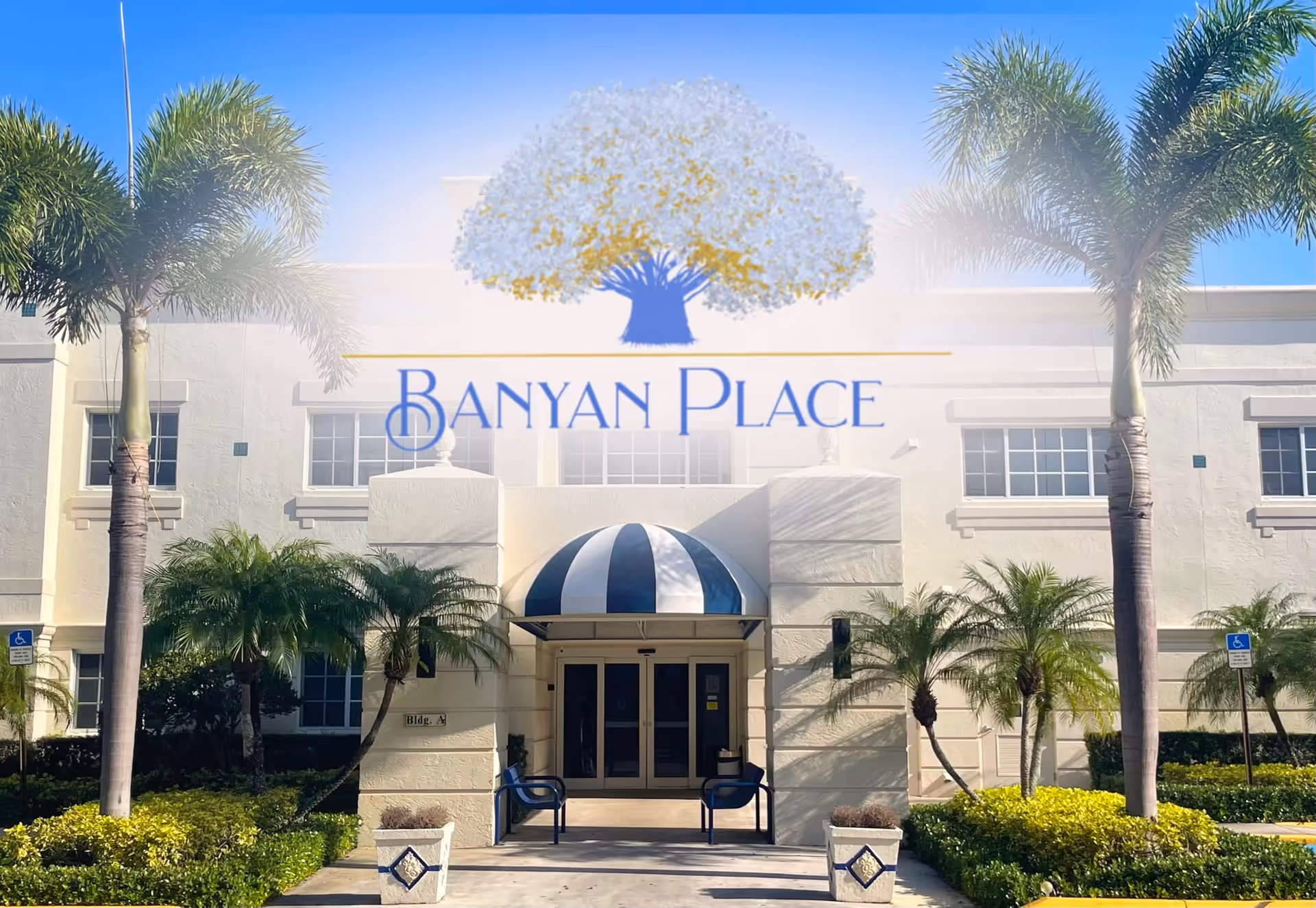 Front exterior view of Banyan Place building entrance with a blue and white striped awning, two blue benches, palm trees, and landscaped greenery under a clear blue sky.