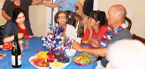 A group of people sitting around a dining table decorated with a blue tablecloth and patriotic-themed centerpiece. The table has plates of fruit, a bottle of wine, and other food items. The people appear to be engaged in conversation and enjoying a meal together.