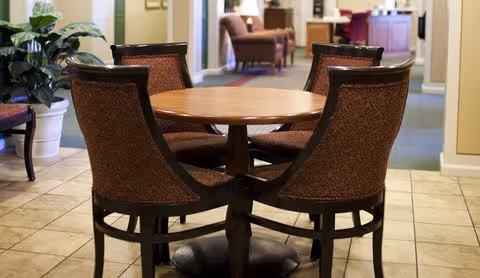 A round wooden table surrounded by four upholstered chairs with a hallway and additional seating visible in the background, inside a senior living facility.