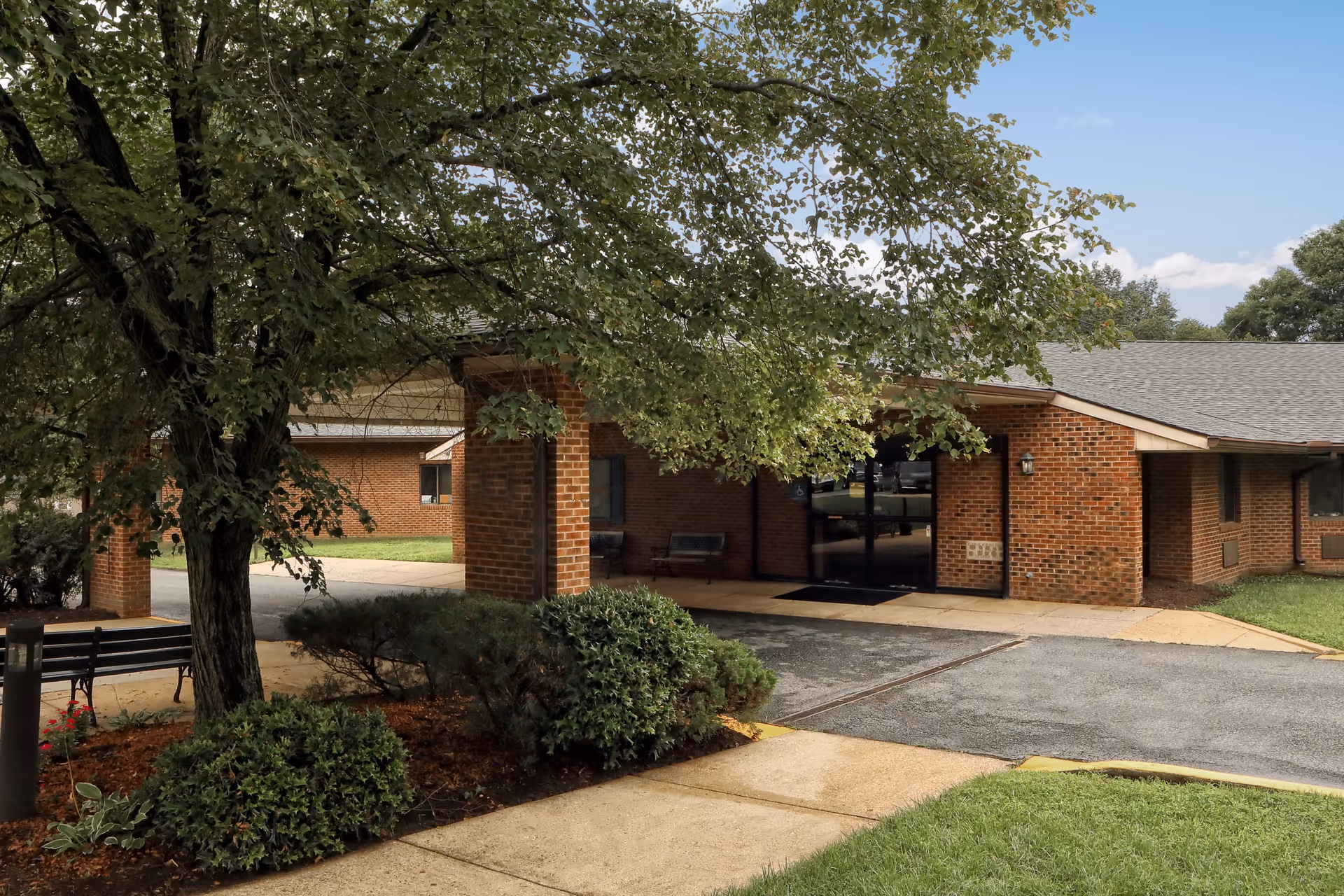 Exterior view of a brick building with a covered entrance, surrounded by green bushes, a large tree, and a well-maintained lawn under a clear blue sky.